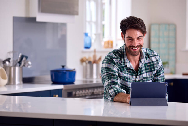 Man Using Digital Tablet In Kitchen To Work From Home Book Holiday Or Shop - Foto, Imagem