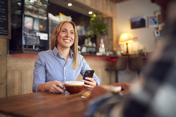 Couple Sitting At Table In Coffee Shop Looking At Mobile Phone Together - Photo, Image