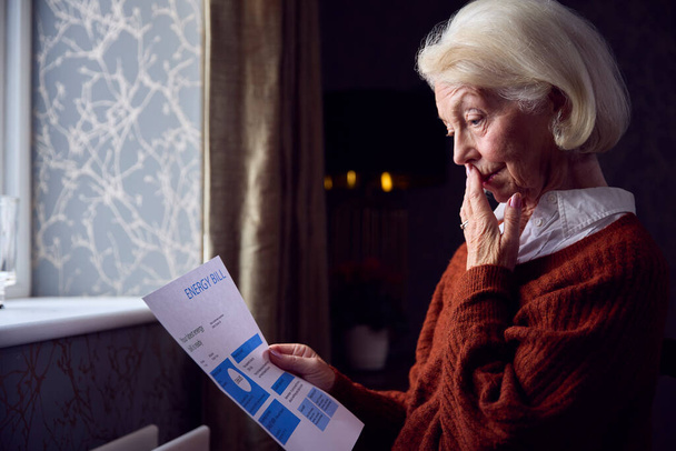 Senior Woman With USA Energy Bill Trying To Keep Warm By Radiator In Cost Of Living Energy Crisis - Фото, изображение