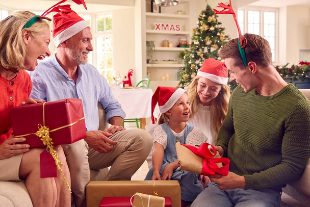Multi-Generation Family Celebrate Christmas At Home Wearing Santa Hats And Antlers Opening Presents - Foto, Bild