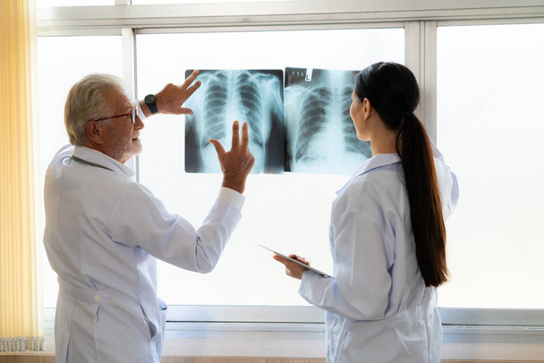 In a hospital sterile room, two professional radiographers hold and examine a radiograph for medical xray diagnosis. Novice doctor seeks advice on a patients condition from experienced older doctor. - Foto, Imagem