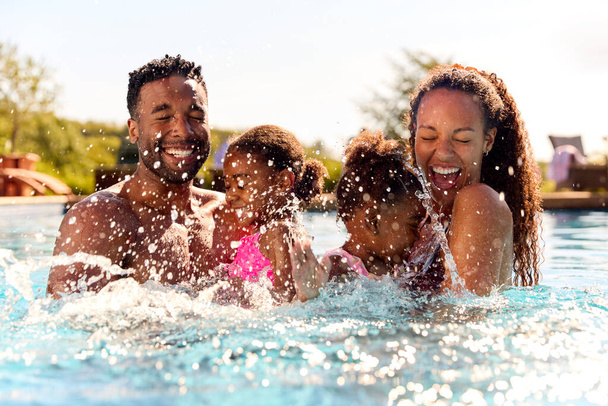 Family On Summer Holiday With Two Girls Being Held In Swimming Pool By Parents And Splashing - Foto, immagini