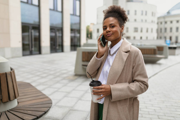 una donna americana adulta in abiti da lavoro parla al telefono con una tazza di caffè in mano sullo sfondo di un edificio per uffici. - Foto, immagini