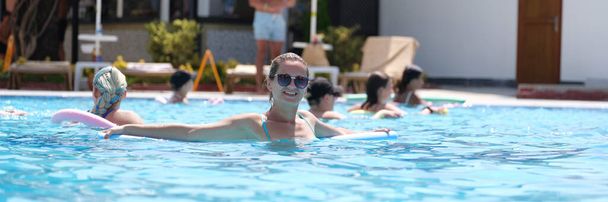 Young woman in sunglasses swims with aqua noodles in pool. Aqua aerobics concept - Foto, afbeelding