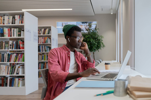 Focalisé jeune enseignant homme afro-américain tuteur regardant ordinateur portable se préparant pour la classe en ligne dans la bibliothèque, gars noir concentré en utilisant l'ordinateur pour l'enseignement ou l'apprentissage. Concept d'enseignement à distance - Photo, image