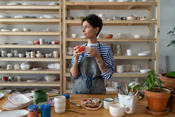 Woman ceramist drinks tea with bun in ceramics studio. Coffee break after pottery master class or work in handicraft shop. Female artist stands against shelving with handmade utensil - Photo, Image