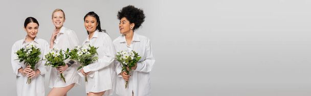 young and pretty multiethnic women in white shirts posing with bouquets isolated on grey, banner - Foto, afbeelding