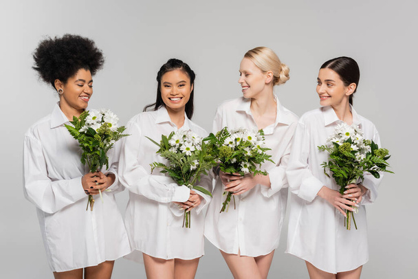 young interracial women in white shirts holding flowers and smiling isolated on grey - Photo, image