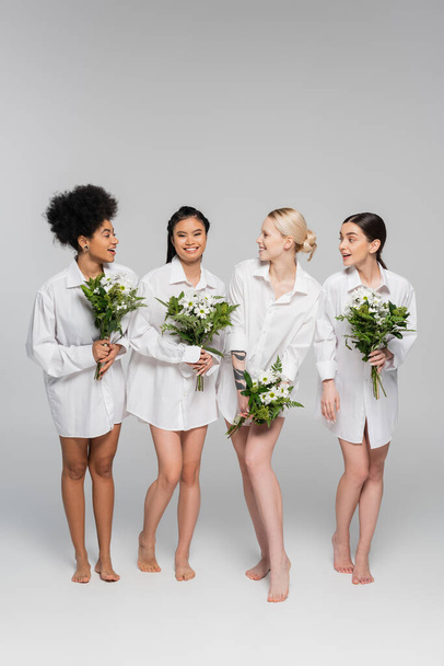 full length of barefoot multicultural women in white shirts holding bouquets on grey background - Foto, immagini
