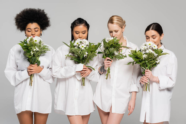  charming multiethnic women with closed eyes enjoying flavor of aromatic flowers isolated on grey - Φωτογραφία, εικόνα