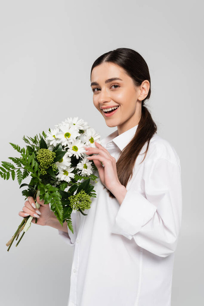 heureuse femme brune en chemise tenant bouquet de fleurs blanches avec des feuilles vertes isolées sur gris - Photo, image