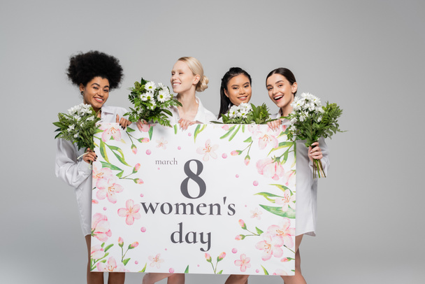 młode międzyrasowe kobiety uśmiechnięte w pobliżu kwiatów i ogromne kobiety dzień kartka okolicznościowa odizolowany na szary - Zdjęcie, obraz