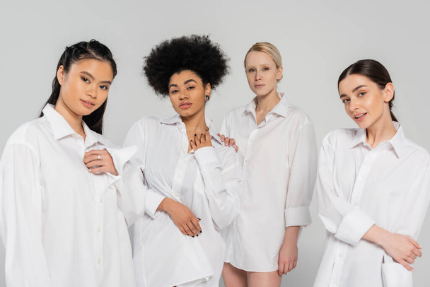 young multiethnic women looking at camera while posing in white shirts isolated on grey - Foto, Imagen