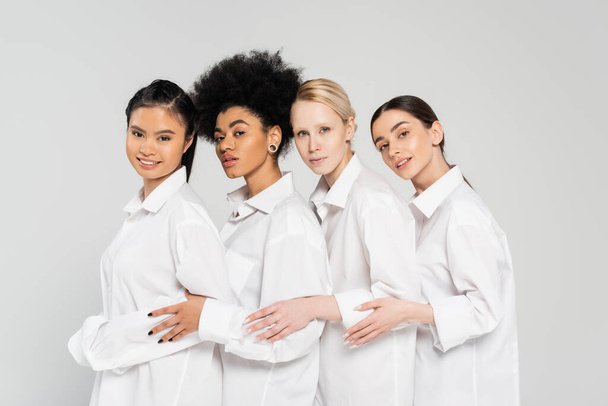 jóvenes mujeres multiétnicas mirando a la cámara mientras posan en camisas blancas aisladas en gris - Foto, Imagen