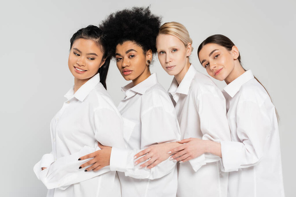 positive and sensual multicultural women looking at camera while posing in white shirts isolated on grey - Photo, image