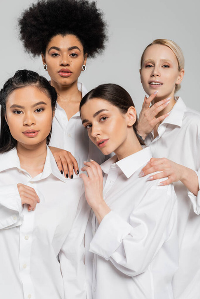 pretty multiethnic women wearing white shirts and looking at camera isolated on grey - 写真・画像