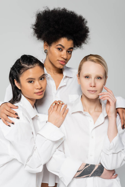 encaracolado afro-americano mulher abraçando ombros de jovens modelos multiétnicos em camisas brancas isoladas em cinza - Foto, Imagem