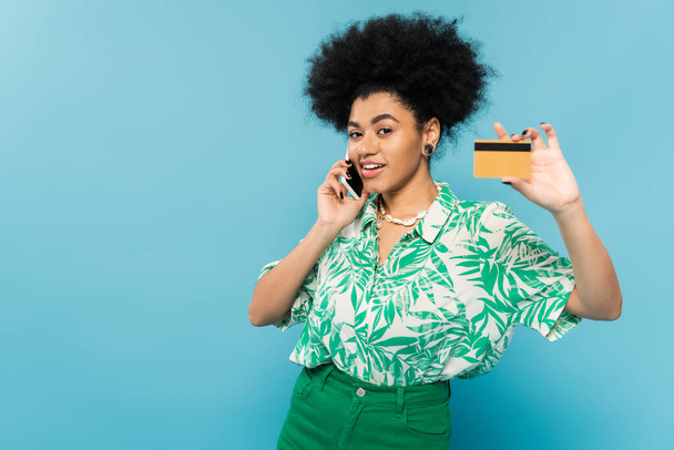 trendy african american woman showing credit and talking on cellphone while smiling at camera isolated on blue - 写真・画像