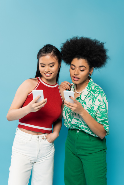 interracial women in stylish clothes looking at mobile phones isolated on blue - Photo, image