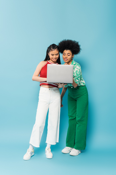 comprimento total de mulher multiétnica jovem em roupas da moda sorrindo enquanto olha para o laptop no fundo azul - Foto, Imagem