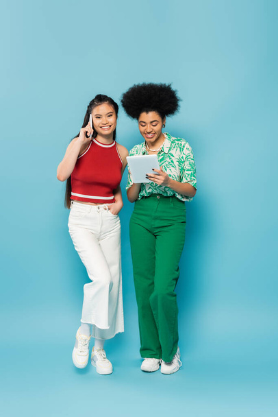 Afro-Amerikan arkadaşının yanında akıllı telefondan konuşan mutlu Asyalı kadın mavi arka planda dijital tablet kullanıyor. - Fotoğraf, Görsel