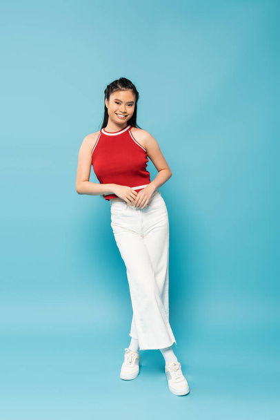 full length of smiling asian woman in red top and white pants standing on blue background - Foto, Imagen