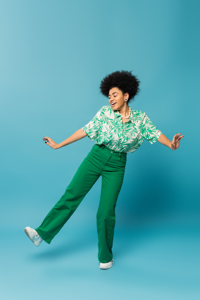 comprimento total de mulher americana africana alegre em folha impressão blusa e calças verdes posando no fundo azul - Foto, Imagem