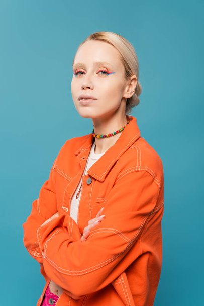 stylish blonde woman in bright orange jacket posing with crossed arms isolated on blue - Foto, Bild