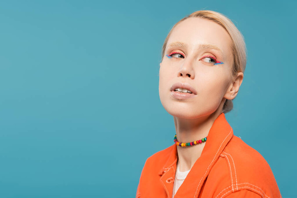 portrait of pretty blonde woman in colorful beads and bright orange jacket looking away isolated on blue - Photo, image