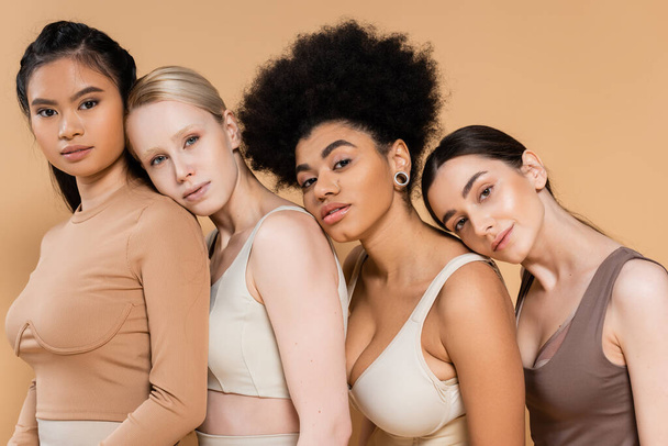 jóvenes mujeres multiétnicas en ropa interior apoyadas una sobre la otra y mirando a la cámara aislada en beige - Foto, Imagen