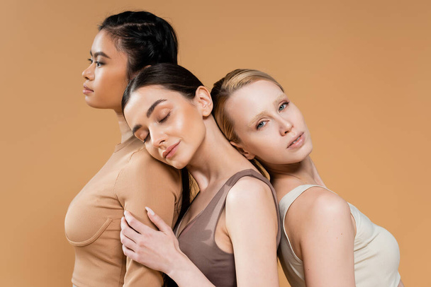 young brunette model posing with closed eyes near interracial women in underwear isolated on beige - Foto, afbeelding