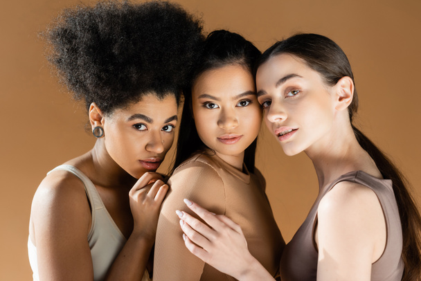 junge und sinnliche multiethnische Frauen in Unterwäsche, die vereinzelt in beige in die Kamera schauen - Foto, Bild