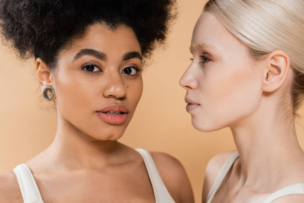 portrait of pretty african american woman looking at camera near young blonde model isolated on beige - Foto, imagen