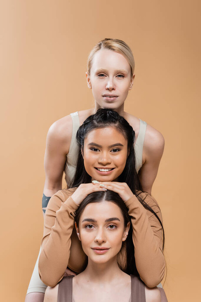 modelos multiculturales positivos en ropa interior sonriendo a la cámara mientras posan aislados en beige - Foto, Imagen
