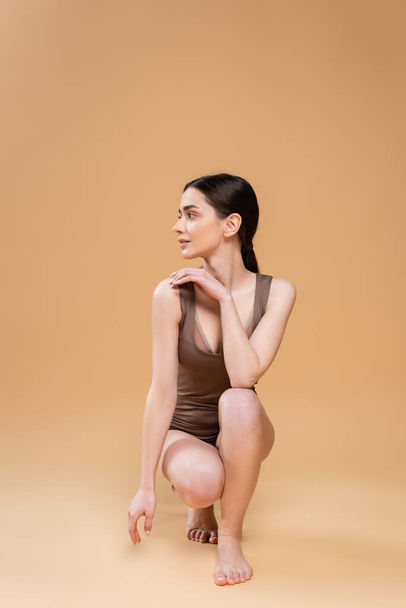 full length of young barefoot woman in underwear looking away while sitting on haunches on beige background - Zdjęcie, obraz