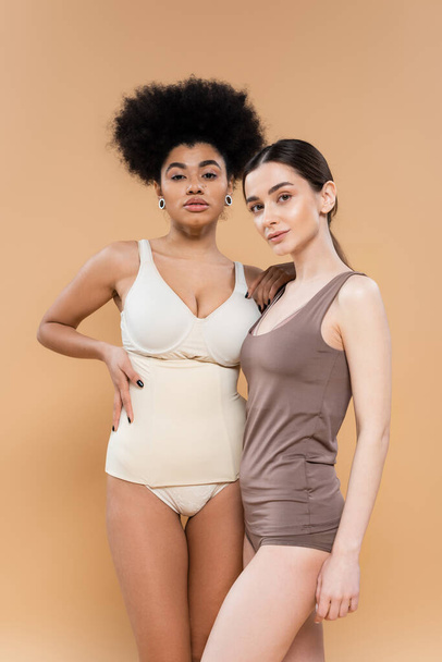 pretty african american model in lingerie posing with hand on waist near young brunette woman isolated on beige - Fotografie, Obrázek