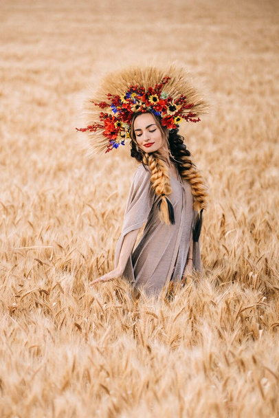 Mulher jovem ucraniana bonita de pé sozinho em um campo de trigo amarelo. A morena olha para a câmera abraçando as espiguetas de trigo. Paz feliz Ucrânia. - Foto, Imagem