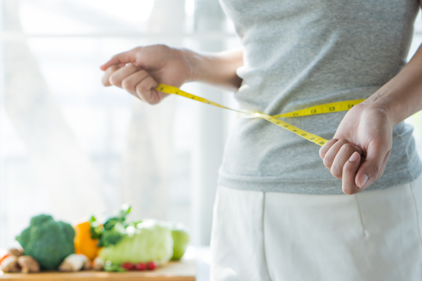 Eat good food for good shape concepts. Woman measuring her body by measure tape have a vegetables on the table as background. Girl checking her waist size down to follow up diet session result. - Foto, immagini