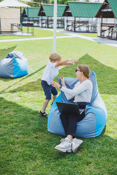 Mother with laptop, her son playing outdoors on green lawn. Rest and work with a child in the park - Fotoğraf, Görsel