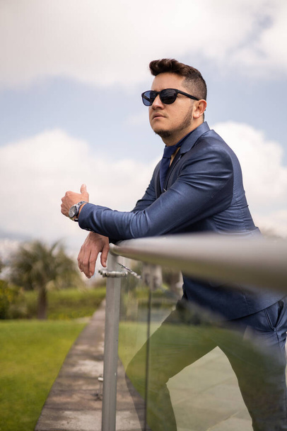 male model resting leaning against a handrail wears a formal suit and sunglasses, young man with short hair, lifestyle and fashion outdoors - Foto, imagen