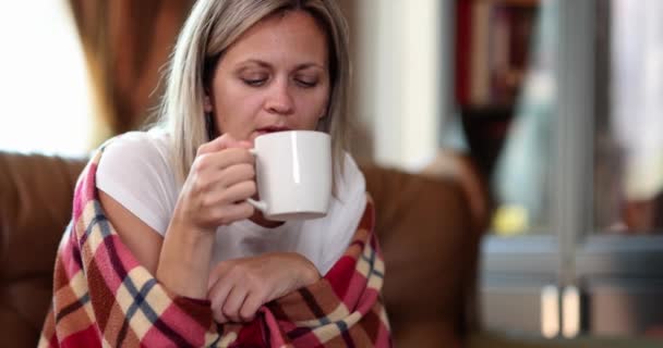 Portrait of sick woman sitting on couch wrapped in blanket and drinking hot tea. Cold flu and pneumonia - Materiaali, video