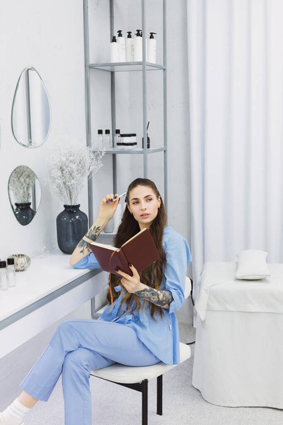 This photo shows a cosmetic dermatologist taking notes at her desk, surrounded by plants, books, and accessories. Her blue coat and professional focus convey a sense of clinical precision. - Foto, imagen