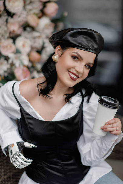 Belleza estilo francés discapacitado Mujer con brazo protésico biónico, mano artificial beber café al aire libre en primavera flor interior café. Vertical. Diversidad femenina - Foto, Imagen