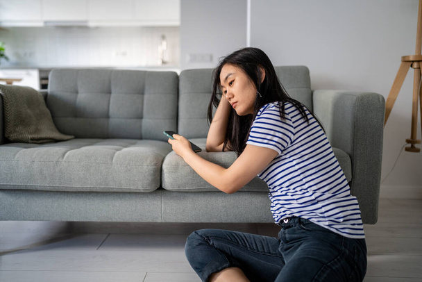 Downcast asian woman looking sadly to device screen reading unpleasant message or waiting for call. Upset Chinese girl using cellphone grudgingly reading news pondering problem considering solutions. - Photo, Image