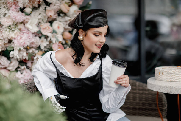 Belleza estilo francés discapacitado Mujer con brazo protésico biónico, mano artificial beber café al aire libre en primavera flor interior café. Vertical. Diversidad femenina - Foto, imagen