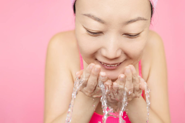 Портрет красивой молодой азиатки для умывания лица брызг воды на розовом фоне. - Фото, изображение