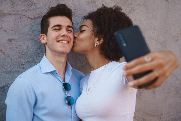 Multiracial couple taking a selfie, a young African American woman with curly hair kissing a Caucasian man on the cheek against a cement wall. Love, interracial, diversity, relationship concepts - 写真・画像