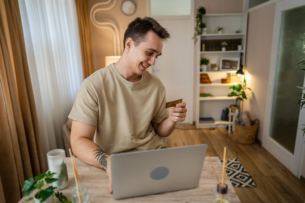 jeden mężczyzna dorosły biały mężczyzna siedzieć w domu szczęśliwy uśmiech na laptop komputer trzymać bank karta kredytowa zakupy koncepcja prawdziwe ludzie kopiować przestrzeń e-commerce - Zdjęcie, obraz