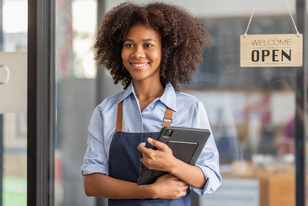 Successful african woman in apron standing coffee shop door. Happy small business owner holding tablet and working. Smiling portrait of SME entrepreneur seller business standing with copy space. - Photo, Image