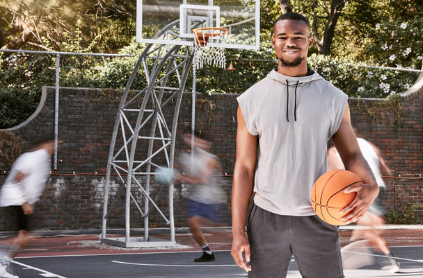 Portrait african american man standing with a basket ball on the court. Handsome male basketball player holding a sports ball while his friends and teammates play and practice in the background. - Photo, Image